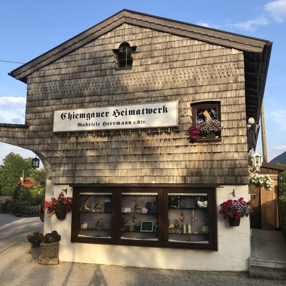Chiemgauer Heimatwerk Gabriele Herrmann in Bernau am Chiemsee Holzverkleidetes Gebäude mit blühenden Blumenkästen und einem Schaufenster.